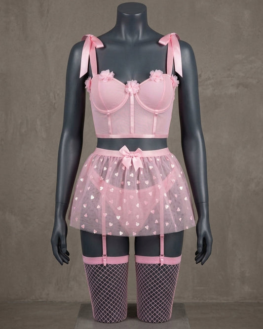 Set de lencería romántica rosa pastel en maniquí gris oscuro, compuesto por top bralette con detalles florales, minifalda de tul translúcido con corazones blancos estampados, ligueros incorporados y medias de red con acabado de encaje rosa.